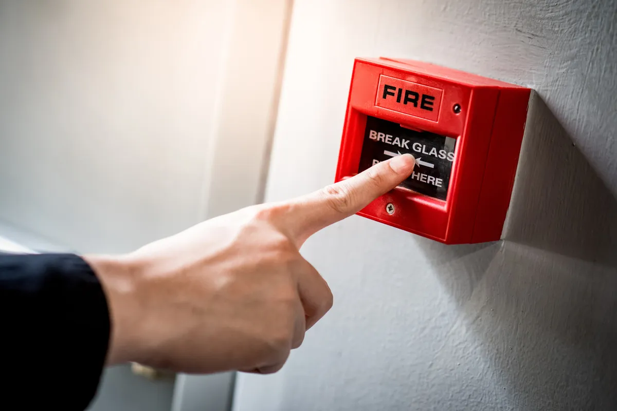 Belys Bau GmbH – Bauunternehmen – Männerhand zeigt auf roten Feuermelder an Betonwand in Bürogebäude